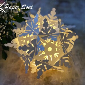Peut inclure: Une lanterne en papier en forme de flocon de neige blanche avec des d&eacute;coupes complexes, illumin&eacute;e de l'int&eacute;rieur, cr&eacute;ant une lueur chaleureuse. La lanterne est faite de papier pli&eacute; et pr&eacute;sente un design d&eacute;licat et tridimensionnel. Le mot "Paper Soul" est visible en arri&egrave;re-plan.
