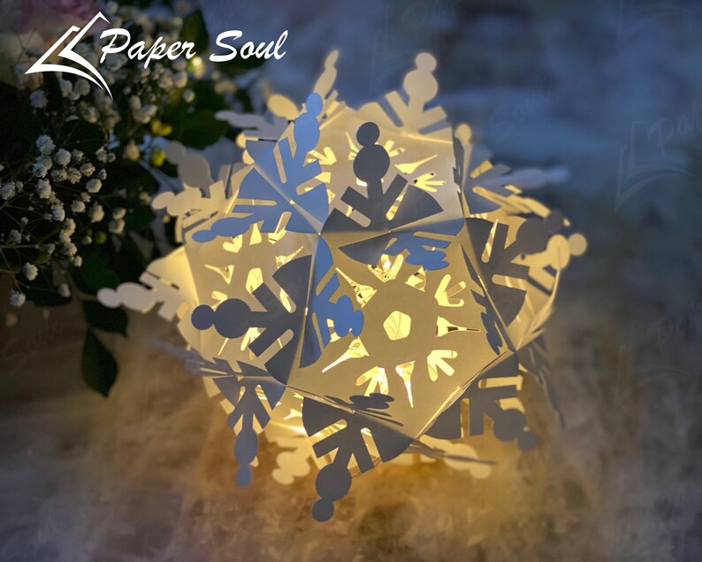 Peut inclure: Une lanterne en papier en forme de flocon de neige blanche avec des d&eacute;coupes complexes, illumin&eacute;e de l'int&eacute;rieur, cr&eacute;ant une lueur chaleureuse. La lanterne est faite de papier pli&eacute; et pr&eacute;sente un design d&eacute;licat et tridimensionnel. Le mot "Paper Soul" est visible en arri&egrave;re-plan.
