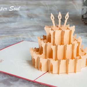 以下が含まれることがあります： ろうそくが立てられた誕生日ケーキが描かれた3Dポップアップカード。カードが開いており、ケーキはカードの中央にあります。カードは白で、赤い縁取りがされています。カードには「ハッピーバースデー」の文字が書かれています。