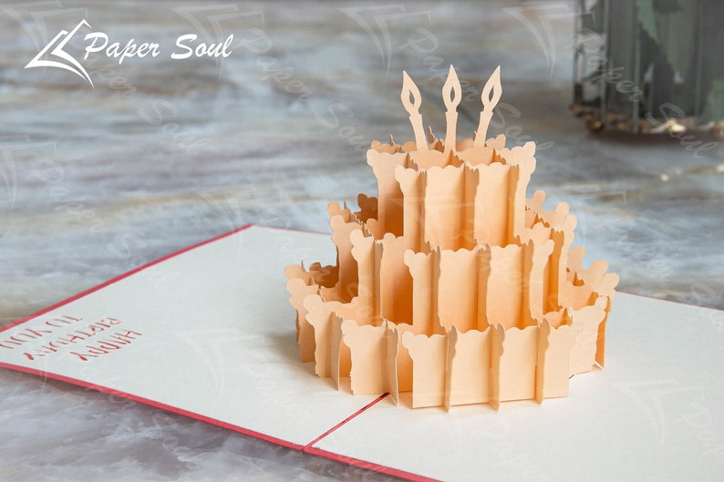 以下が含まれることがあります： ろうそくが立てられた誕生日ケーキが描かれた3Dポップアップカード。カードが開いており、ケーキはカードの中央にあります。カードは白で、赤い縁取りがされています。カードには「ハッピーバースデー」の文字が書かれています。