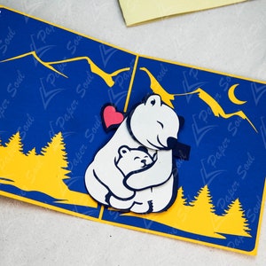 May include: A 3D pop-up card featuring a white polar bear hugging a smaller polar bear. The card has a blue background with yellow mountains and trees. A red heart is visible between the bears.