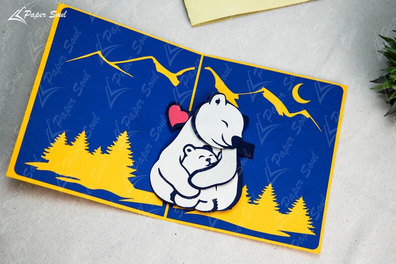 May include: A 3D pop-up card featuring a white polar bear hugging a smaller polar bear. The card has a blue background with yellow mountains and trees. A red heart is visible between the bears.