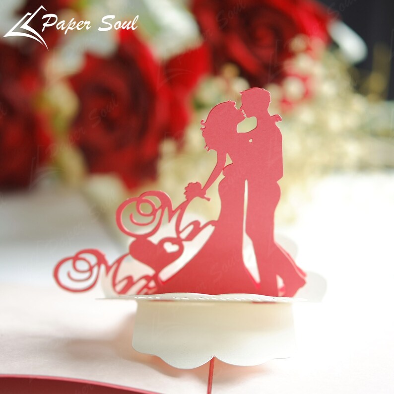 Pode incluir: Um cart&atilde;o pop-up de papel vermelho com uma silhueta de um casal se beijando. O cart&atilde;o &eacute; decorado com um cora&ccedil;&atilde;o e as palavras "Mr & Mrs".