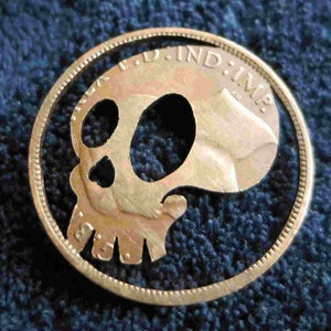 Puede incluir: Una moneda de color dorado con un cráneo recortado en el centro. La moneda tiene la inscripción "F.D. IND: IMP." alrededor del borde.