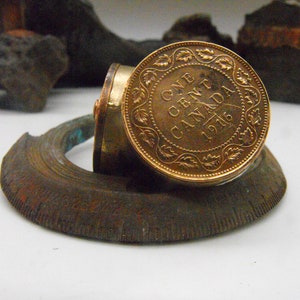 May include: A gold-coloured Canadian coin from 1916, with the inscription "ONE CENT CANADA 1916", is mounted on a circular, brown, metal base.