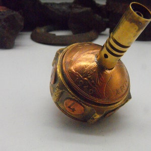 May include: A copper and brass spinning top with a coin on the bottom. The top has a brass tube on top and a coin on each side.