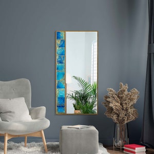 May include: A tall, rectangular mirror with a gold frame. The mirror has a blue and gold patterned border on the left side. The mirror is hanging on a grey wall.