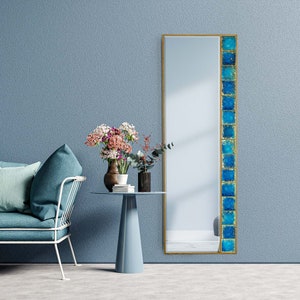 May include: A full-length mirror with a gold frame and a vertical strip of blue and gold tiles on the right side. The mirror is reflecting a room with a sofa, a side table, and flowers.