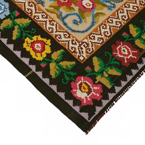 May include: Close-up of a corner of a colorful, patterned rug. The rug features a black border with floral designs in red, yellow, and pink. The central design has a beige background with floral and scroll patterns.