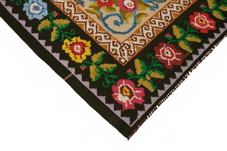 May include: Close-up of a corner of a colorful, patterned rug. The rug features a black border with floral designs in red, yellow, and pink. The central design has a beige background with floral and scroll patterns.