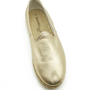 May include: A single gold leather loafer shoe. The shoe has a rounded toe and a flat sole. The insole is tan with the brand name "Svony Handmade Shoes" printed on it. The shoe is size 42.