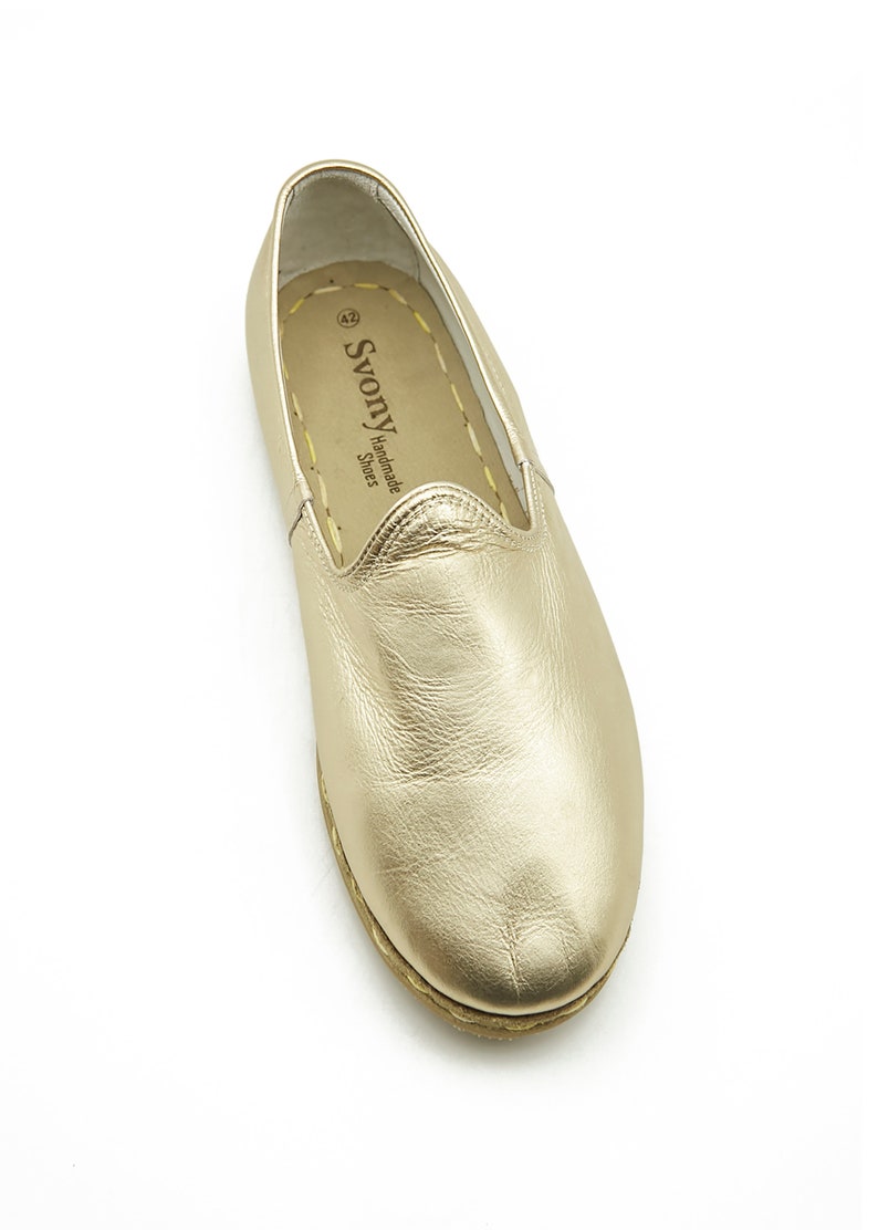 May include: A single gold leather loafer shoe. The shoe has a rounded toe and a flat sole. The insole is tan with the brand name "Svony Handmade Shoes" printed on it. The shoe is size 42.