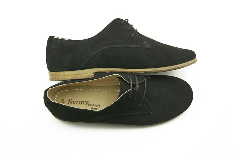 May include: A pair of black suede lace-up shoes with a brown leather sole. The shoes are handmade and have the brand name "Svonya" printed on the insole.