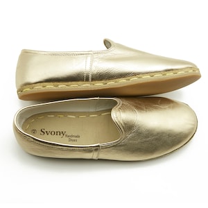 May include: A pair of gold leather slip-on shoes with a rounded toe and a low heel. The shoes are presented on a white background. The brand name "Svony Handmade Shoes" is visible inside one of the shoes.
