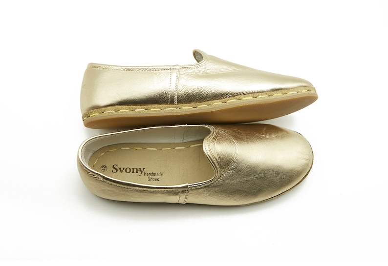 May include: A pair of gold leather slip-on shoes with a rounded toe and a low heel. The shoes are presented on a white background. The brand name "Svony Handmade Shoes" is visible inside one of the shoes.