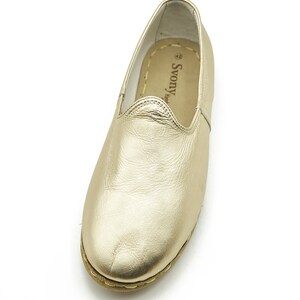 May include: Gold-coloured leather slip-on shoe. The shoe has a rounded toe and a stitched sole. The interior is a light tan colour with the brand name "Svony Hand" printed on the insole. The shoe is designed for comfort and style.