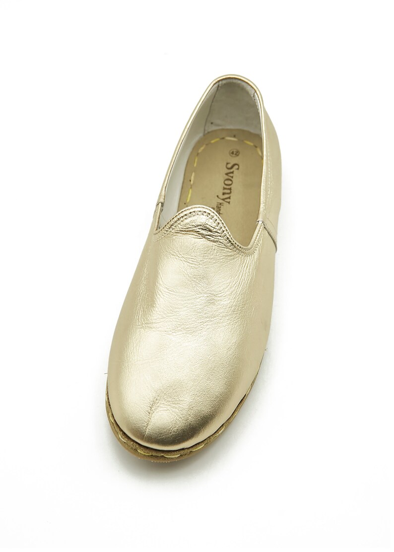 May include: Gold-coloured leather slip-on shoe. The shoe has a rounded toe and a stitched sole. The interior is a light tan colour with the brand name "Svony Hand" printed on the insole. The shoe is designed for comfort and style.