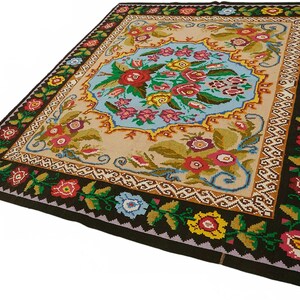 May include: A rectangular rug with a central floral design in a light blue oval shape. The rug features a beige background with a dark brown border adorned with colorful flowers and green leaves. The rug's dimensions are not visible.