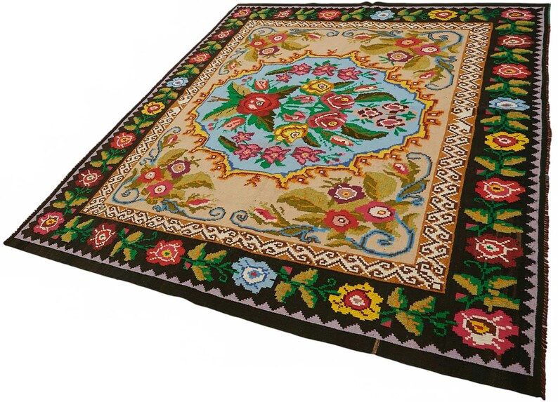 May include: A rectangular rug with a central floral design in a light blue oval shape. The rug features a beige background with a dark brown border adorned with colorful flowers and green leaves. The rug's dimensions are not visible.