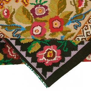May include: A colorful, hand-woven rug with a floral design. The rug features a beige background with red, pink, and purple flowers, green leaves, and blue accents. The border is black with a repeating floral pattern.