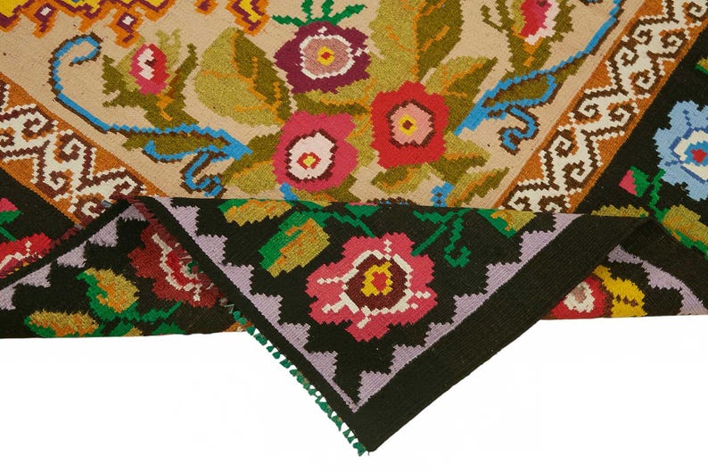 May include: A colorful, hand-woven rug with a floral design. The rug features a beige background with red, pink, and purple flowers, green leaves, and blue accents. The border is black with a repeating floral pattern.