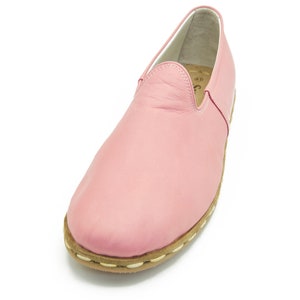 May include: A pair of pink leather slip-on shoes with a white stitching detail around the sole.