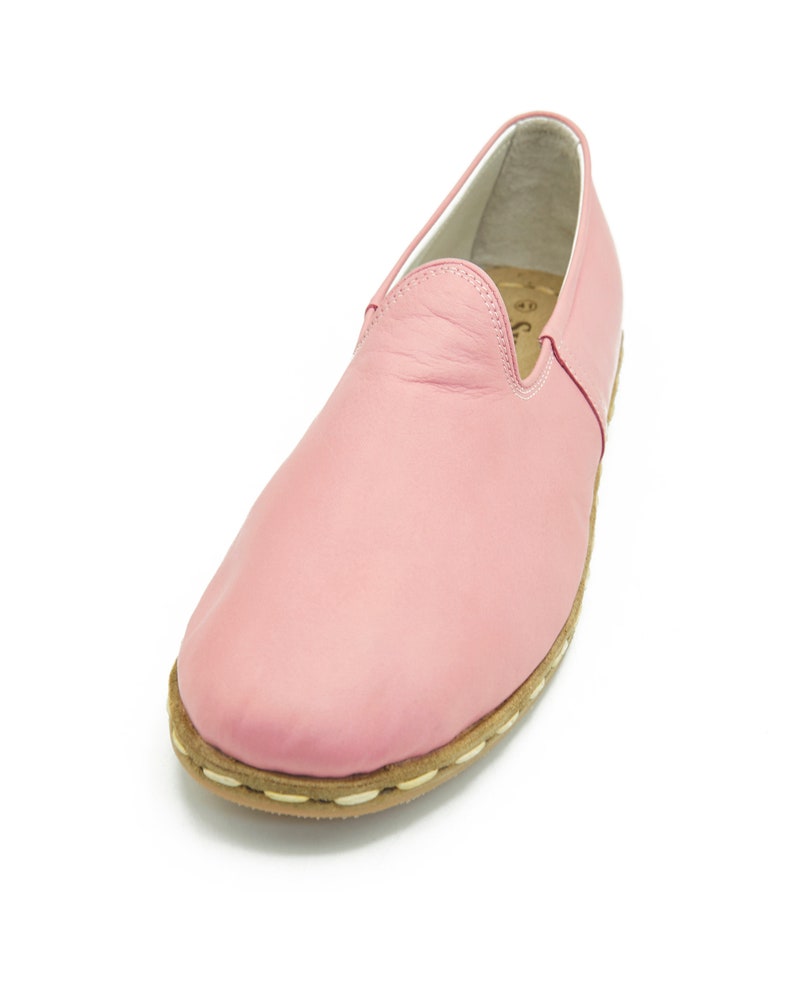 May include: A pair of pink leather slip-on shoes with a white stitching detail around the sole.
