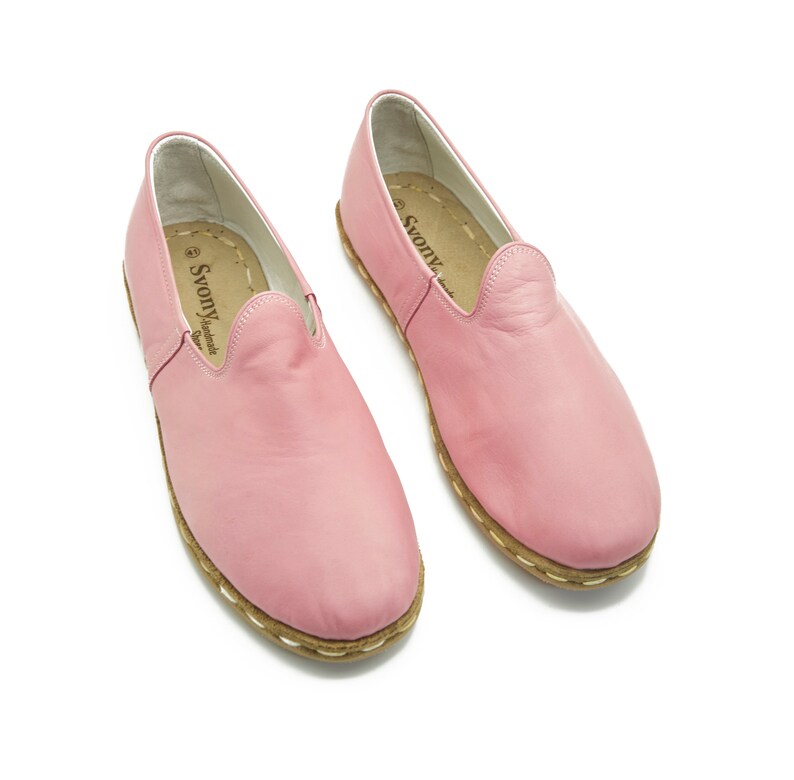 May include: A pair of pink leather slip-on shoes with a white stitched trim around the sole. The shoes have a label that reads "SvonY Handmade Shoes".