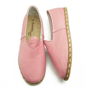 May include: A pair of pink leather slip-on shoes with a brown stitched sole. The shoes have a label inside that reads "Svonya Handmade Shoes".