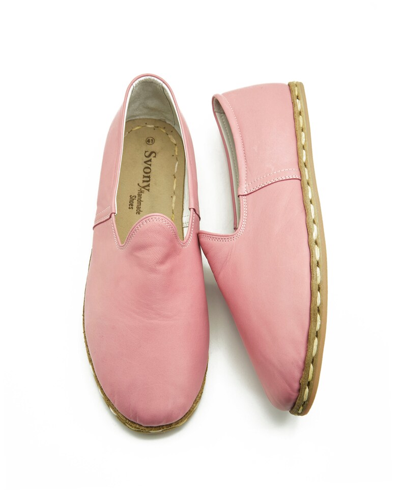 May include: A pair of pink leather slip-on shoes with a brown stitched sole. The shoes have a label inside that reads "Svonya Handmade Shoes".