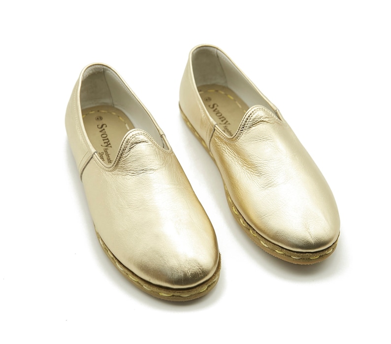 May include: A pair of gold leather slip-on shoes. The shoes have a rounded toe and a low profile. The soles are light brown with visible stitching. The brand name "Svony" is visible on the insole.