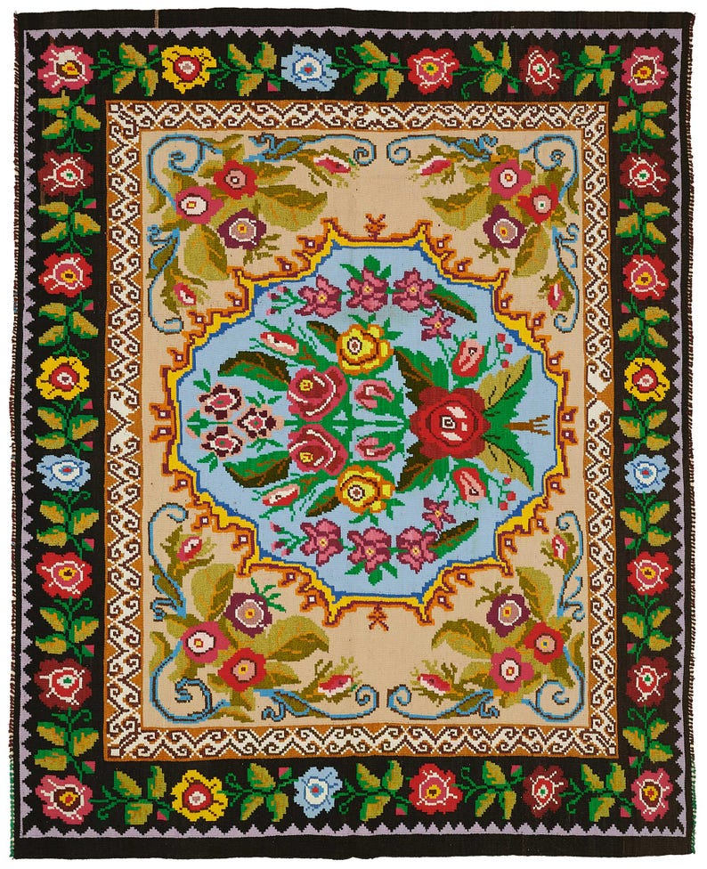 May include: A rectangular, hand-woven rug with a floral design. The rug features a central light blue medallion with a bouquet of red, pink, and yellow flowers. The border is black with repeating floral patterns in red, yellow, and blue. The rug's dimensions are approximately 60 inches by 40 inches.