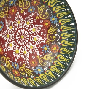K&ouml;nnte beinhalten: Eine runde Keramiksch&uuml;ssel mit einem dunkelgr&uuml;nen Rand und einem farbenfrohen Blumendesign. Die Sch&uuml;ssel hat ein zentrales Motiv aus wei&szlig;en Bl&uuml;ten mit gelben Zentren, umgeben von kleineren roten, gelben und blauen Bl&uuml;ten. Der Rand ist mit gelben und wei&szlig;en stilisierten Flammen verziert.