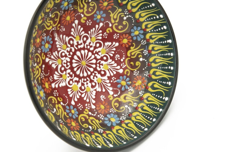 K&ouml;nnte beinhalten: Eine runde Keramiksch&uuml;ssel mit einem dunkelgr&uuml;nen Rand und einem farbenfrohen Blumendesign. Die Sch&uuml;ssel hat ein zentrales Motiv aus wei&szlig;en Bl&uuml;ten mit gelben Zentren, umgeben von kleineren roten, gelben und blauen Bl&uuml;ten. Der Rand ist mit gelben und wei&szlig;en stilisierten Flammen verziert.