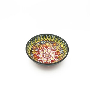 K&ouml;nnte beinhalten: Eine kleine, rote Keramiksch&uuml;ssel mit schwarzem Rand und aufwendigen floralen und geometrischen Mustern in Gelb, Blau und Wei&szlig;.