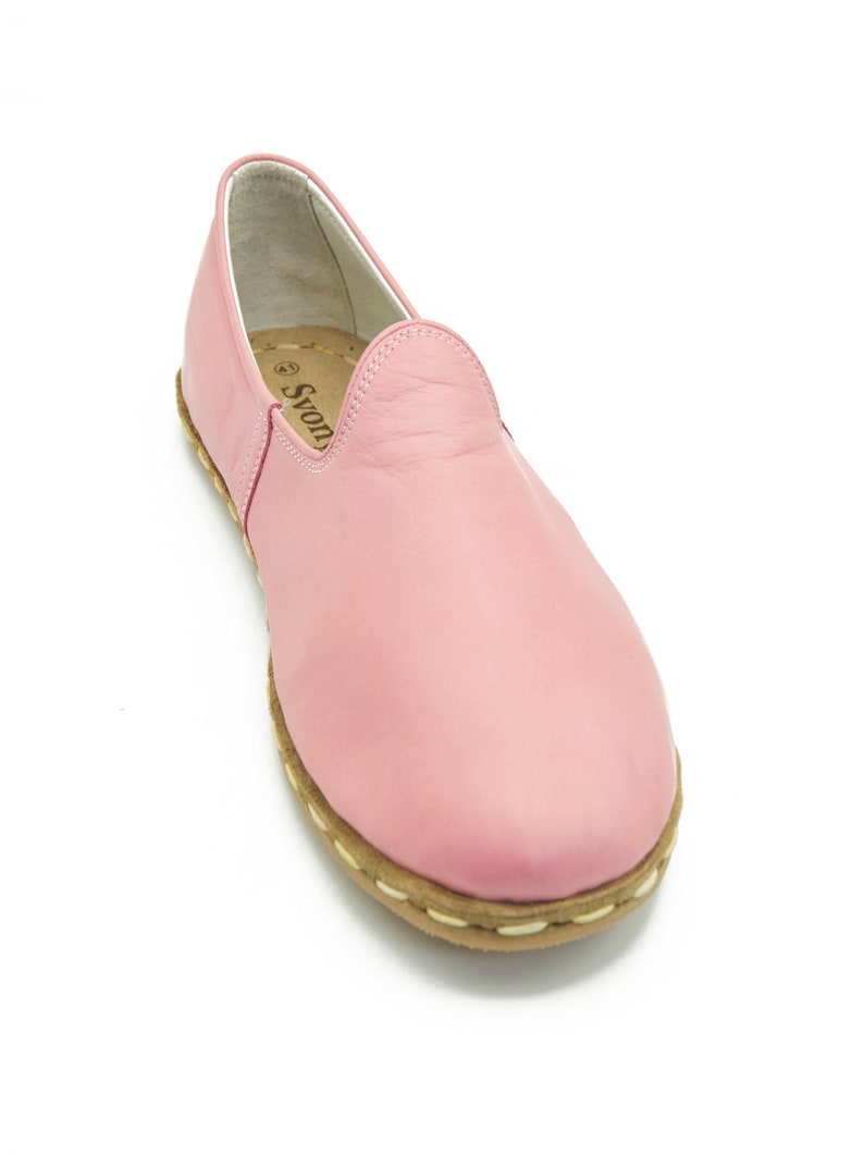 May include: A pair of pink leather slip-on shoes with a white stitched trim around the sole. The shoes have a brand name "Swon" on the inside.