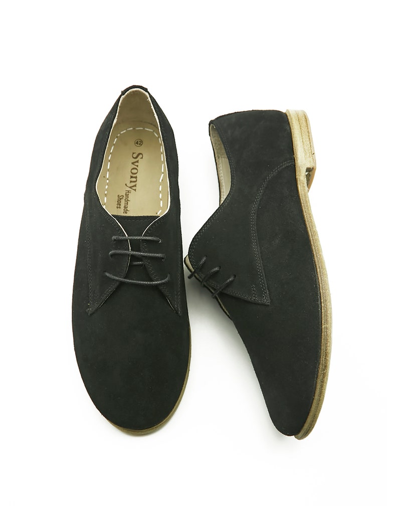 May include: A pair of black suede lace-up shoes with a brown sole. The shoes are handmade by Svonya Shoes.