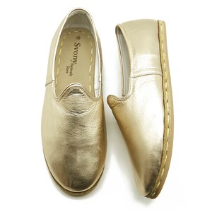 May include: Two gold-coloured leather slip-on shoes. The shoes have a rounded toe and a stitched sole. The interior of one shoe is visible, with the brand name "Svony Handmade Shoes" printed on the insole.