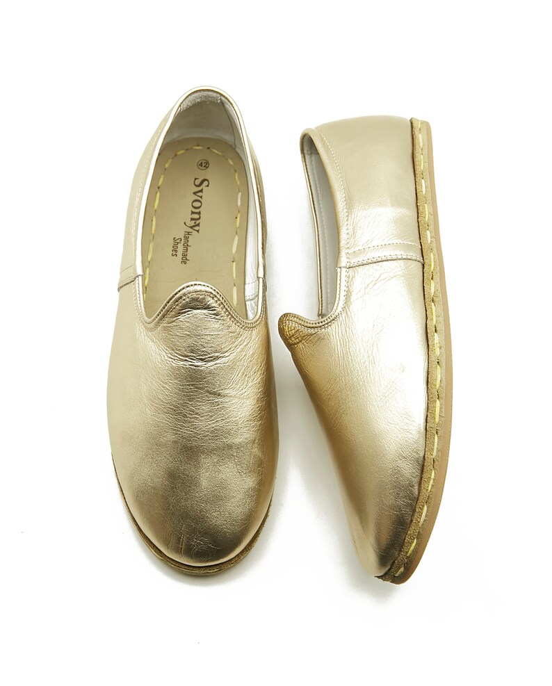 May include: Two gold-coloured leather slip-on shoes. The shoes have a rounded toe and a stitched sole. The interior of one shoe is visible, with the brand name "Svony Handmade Shoes" printed on the insole.