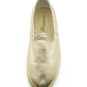May include: A gold leather slip-on shoe. The shoe has a rounded toe and a stitched sole. The brand name "Svony Handmade Shoes" is visible on the side. The shoe is size 42.
