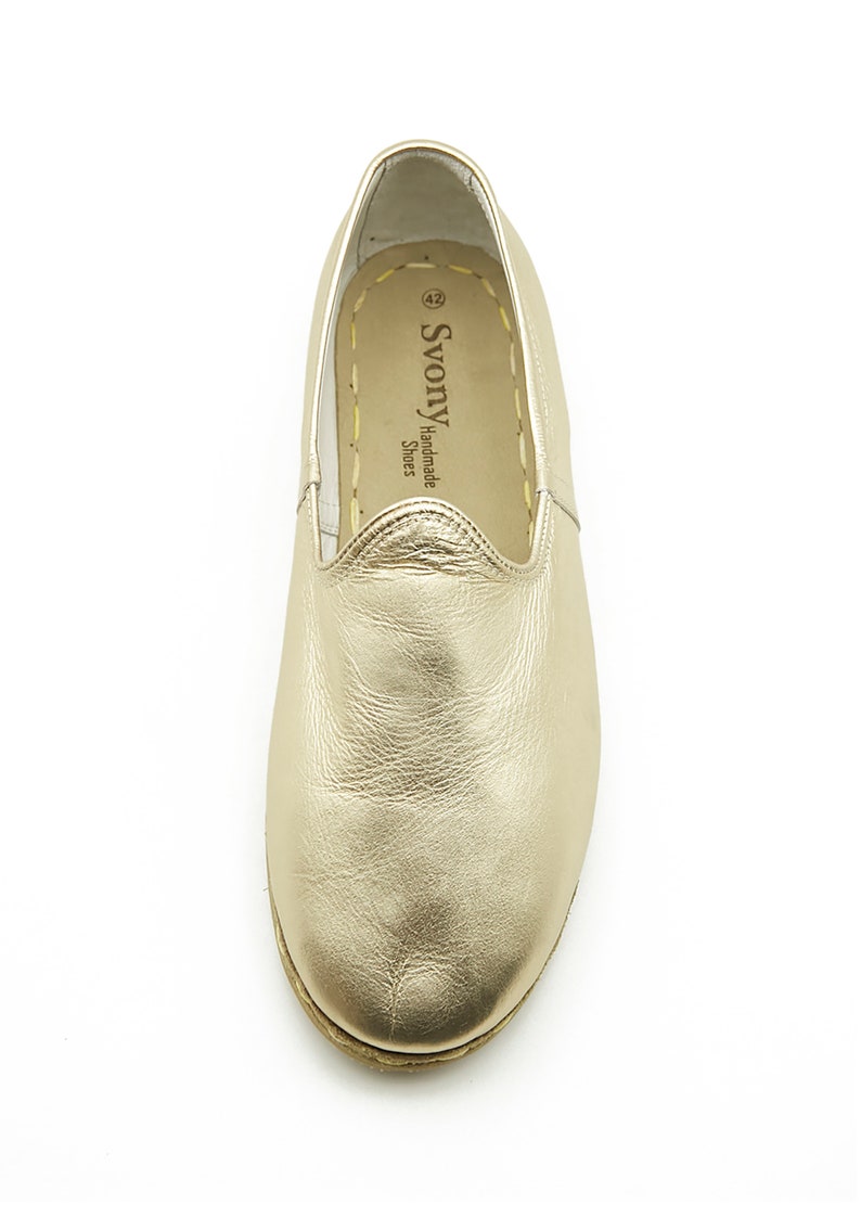May include: A gold leather slip-on shoe. The shoe has a rounded toe and a stitched sole. The brand name "Svony Handmade Shoes" is visible on the side. The shoe is size 42.