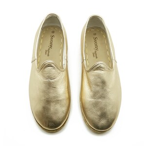 May include: A pair of gold leather slip-on shoes. The shoes have a rounded toe and stitched soles. The brand name "Svony Handmade Shoes" is printed on the insole. The shoes are presented on a white background.