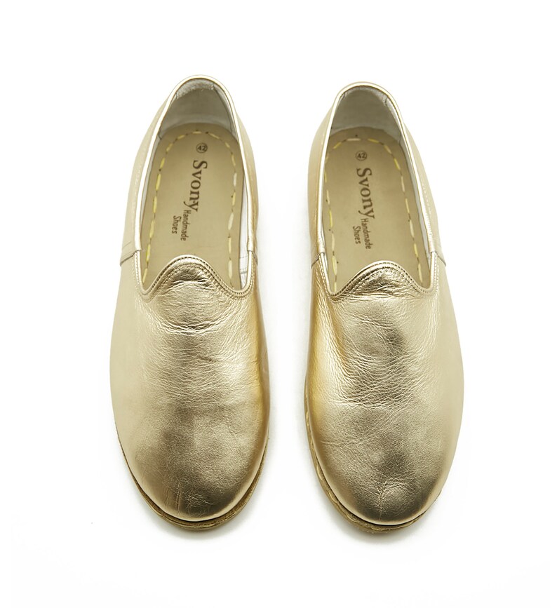 May include: A pair of gold leather slip-on shoes. The shoes have a rounded toe and stitched soles. The brand name "Svony Handmade Shoes" is printed on the insole. The shoes are presented on a white background.
