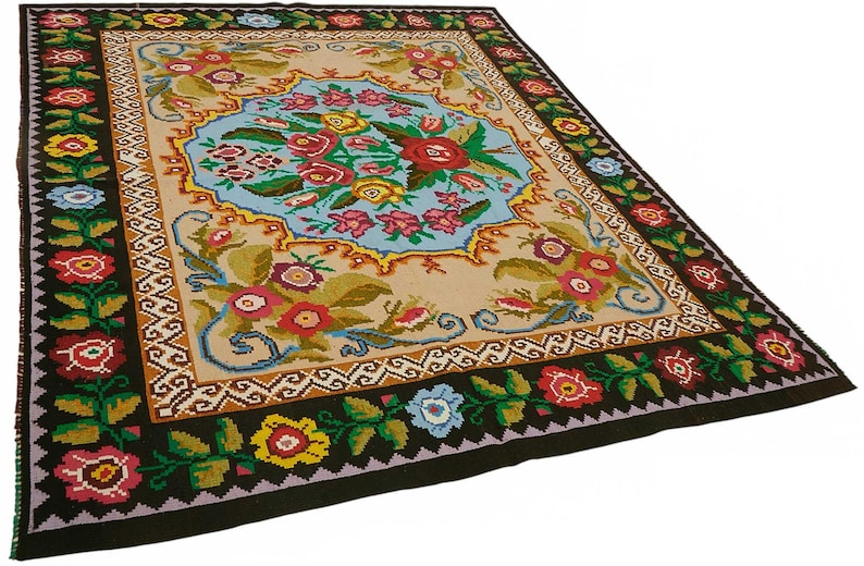 May include: A rectangular, hand-woven rug with a floral design. The rug features a central light blue medallion with a bouquet of red, yellow, and pink flowers. The border is black with colorful floral accents, and the rug measures approximately 60 x 90 inches.