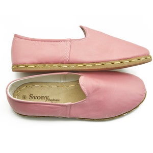 May include: A pair of pink leather slip-on shoes with a brown sole and a stitched seam around the edge. The shoes have a label that reads "Svonya Handmade".
