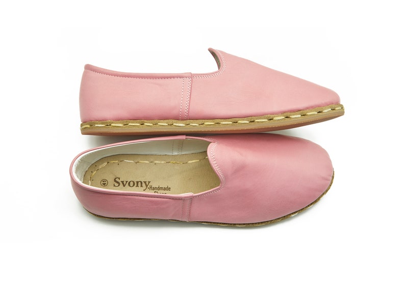 May include: A pair of pink leather slip-on shoes with a brown sole and a stitched seam around the edge. The shoes have a label that reads "Svonya Handmade".