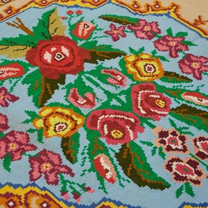 May include: A detailed close-up of a floral rug with a light blue center. The rug features a bouquet of red, pink, and yellow flowers with green leaves. The border is a mix of yellow, orange, and blue geometric patterns. The rug is a decorative textile.
