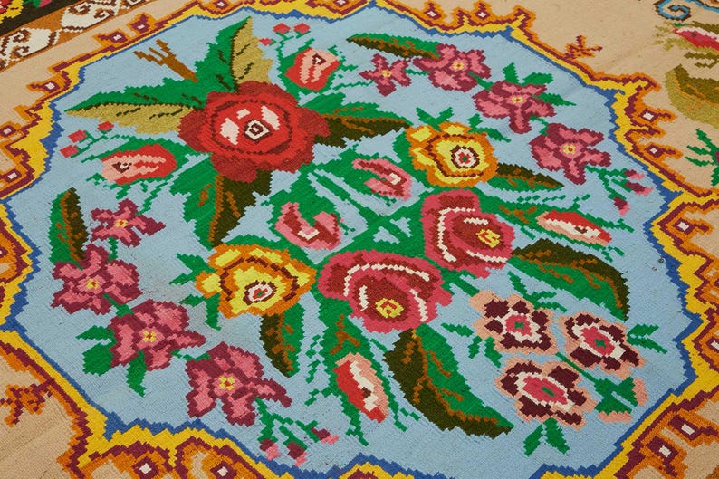 May include: A detailed close-up of a floral rug with a light blue center. The rug features a bouquet of red, pink, and yellow flowers with green leaves. The border is a mix of yellow, orange, and blue geometric patterns. The rug is a decorative textile.