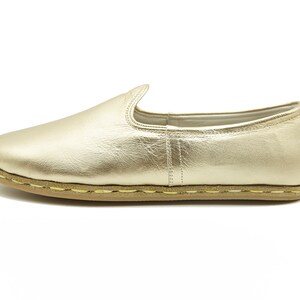 May include: A single gold slip-on shoe. The shoe has a rounded toe and a low profile. The sole is light brown with visible stitching. The upper is a metallic gold material. The shoe is designed for casual wear.