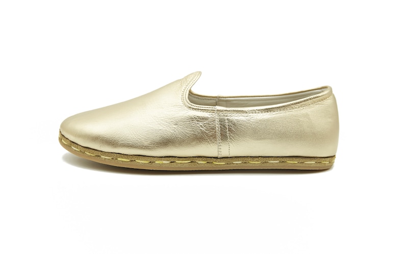 May include: A single gold slip-on shoe. The shoe has a rounded toe and a low profile. The sole is light brown with visible stitching. The upper is a metallic gold material. The shoe is designed for casual wear.